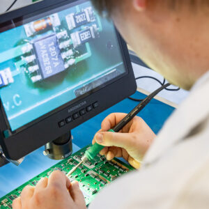 Elektronikfertigung – Person prüft Platine mit Mikroskop und Lötkolben
